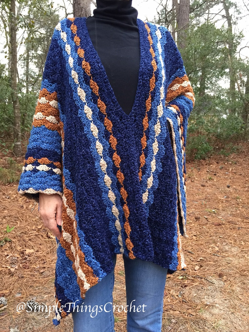 Puede incluir: Un chal de ganchillo hecho a mano, azul, marr&oacute;n y blanco, con cuello en V. El chal presenta un patr&oacute;n texturizado con rayas de varios colores y un ajuste holgado. Las mangas son anchas y el chal se lleva sobre un cuello alto negro y vaqueros azules.
