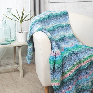 May include: A handmade crochet blanket draped over a white armchair. The blanket features a wave pattern in shades of blue, green, and pink. The text "@SimpleThingsCrochet" is visible in the lower left corner. The blanket is a cozy home decor item.