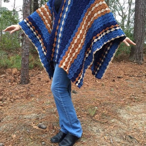 Puede incluir: Un poncho de ganchillo azul, marr&oacute;n y crema. El poncho tiene un patr&oacute;n de rayas con un dise&ntilde;o de cuello en V. La persona de la imagen lleva vaqueros azules y zapatos negros. El texto "@Simple Things Crochet" es visible en la parte inferior de la imagen.