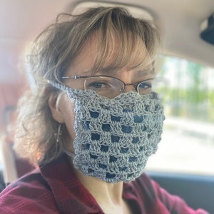 Könnte beinhalten: Nahaufnahme einer Person, die eine graue gehäkelte Gesichtsmaske mit quadratischem Muster trägt. Die Maske bedeckt Nase und Mund und hat ein Band, das um die Ohren geht. Die Person trägt eine Brille und hat kurze, hellbraune Haare. Das Bild ist aus einem leicht niedrigen Winkel aufgenommen.