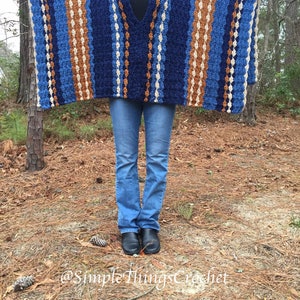 Puede incluir: Un poncho de ganchillo azul, marr&oacute;n y crema con cuello en V. El poncho tiene rayas verticales de diferentes anchos y colores. La persona de la imagen lleva vaqueros azules y botas negras. El fondo es un entorno forestal.