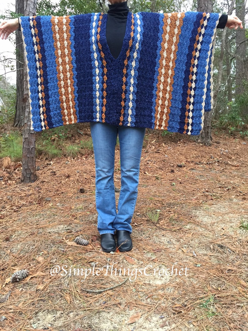 Puede incluir: Un poncho de ganchillo azul, marr&oacute;n y crema con cuello en V. El poncho tiene rayas verticales de diferentes anchos y colores. La persona de la imagen lleva vaqueros azules y botas negras. El fondo es un entorno forestal.