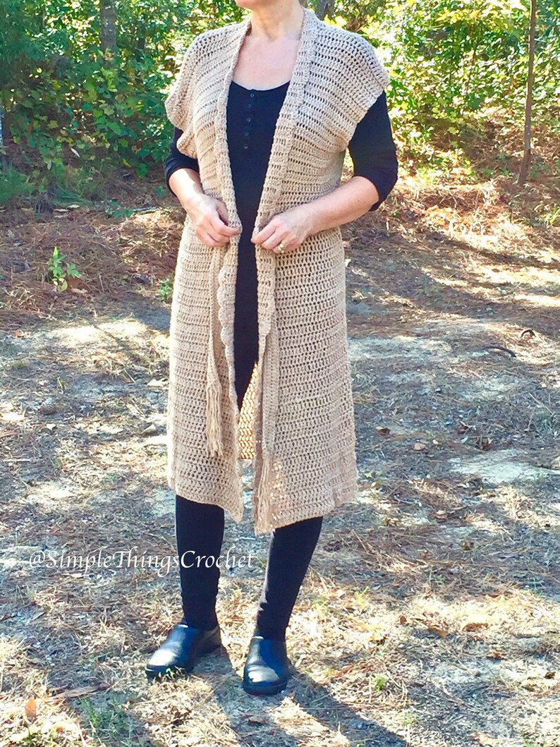 Puede incluir: Un c&aacute;rdigan largo de crochet de color beige con cuello chal y detalles de flecos. El c&aacute;rdigan est&aacute; abierto en la parte delantera y tiene un cierre de corbata. El c&aacute;rdigan se lleva sobre una blusa negra y leggings negros.