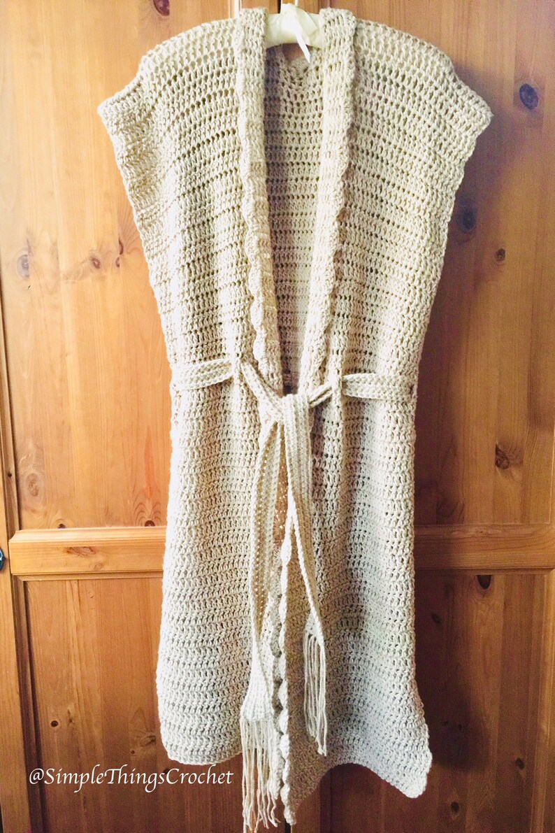 Puede incluir: Un c&aacute;rdigan de crochet beige con un frente largo y abierto y un cintur&oacute;n de lazo. El c&aacute;rdigan tiene un borde de flecos y un dise&ntilde;o de crochet.