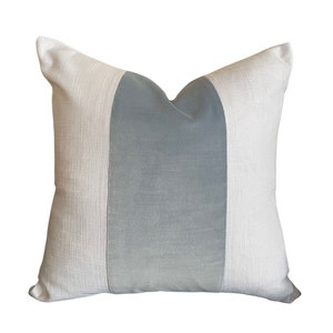 May include: Square decorative pillow with a light gray center panel and off-white side panels. The pillow has a textured appearance and a soft, inviting aesthetic. A simple, yet elegant home decor item.