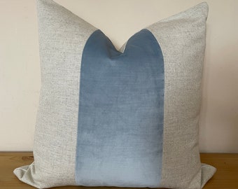 May include: Square decorative pillow with a light gray textured fabric on the sides and a central panel of soft, light blue velvet. The pillow is a neutral color and would complement any home decor.