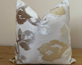 May include: A decorative pillow with a white background and a pattern of abstract shapes in shades of gold, tan, and gray. The pillow has a soft, textured appearance and is likely made of a woven fabric. The pillow is square and appears to be filled.
