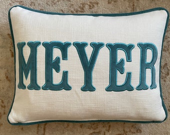 Op de afbeelding: Rechthoekig wit kussen met de naam "MEYER" geborduurd in turquoise letters. Het kussen heeft een turquoise rand en een textuurstof. Een decoratief woonaccessoire.