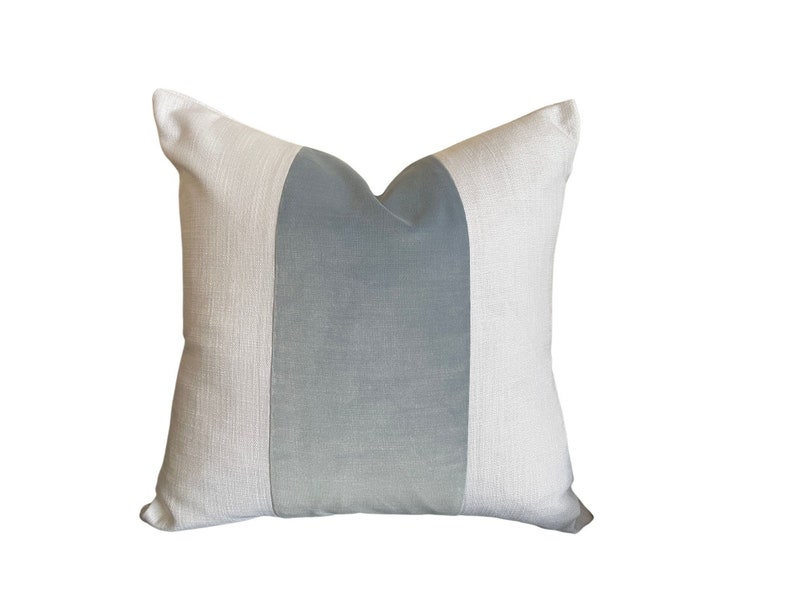 May include: Square decorative pillow with a light gray center panel and off-white side panels. The pillow has a textured appearance and a soft, inviting aesthetic. A simple, yet elegant home decor item.