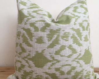 May include: A square decorative pillow with a green and gray abstract pattern. The pillow has a textured appearance and is placed on a wooden surface. The pattern is a repeating design of organic shapes.