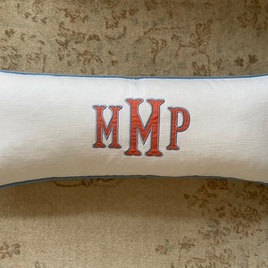 May include: A rectangular white cushion with a blue trim. The cushion displays the embroidered initials "MMP" in orange with a blue outline. The cushion is placed on a patterned beige rug.