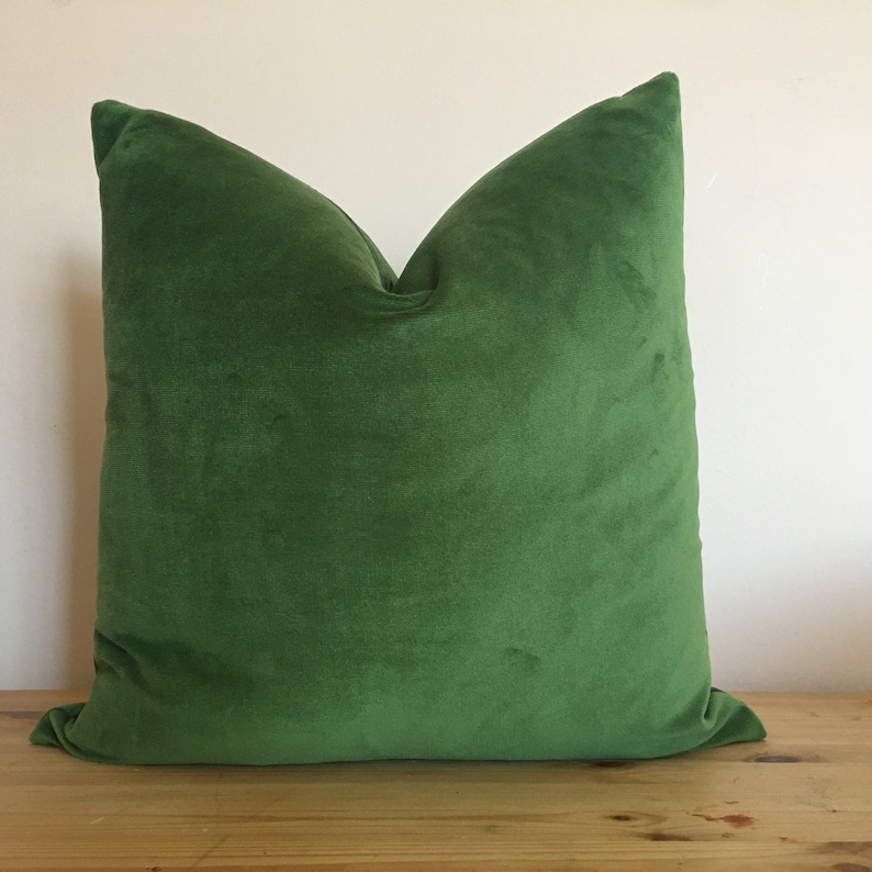 May include: A square, emerald green velvet pillow. The pillow is plush and has a soft, velvety texture. The pillow is resting on a wooden surface against a white wall.