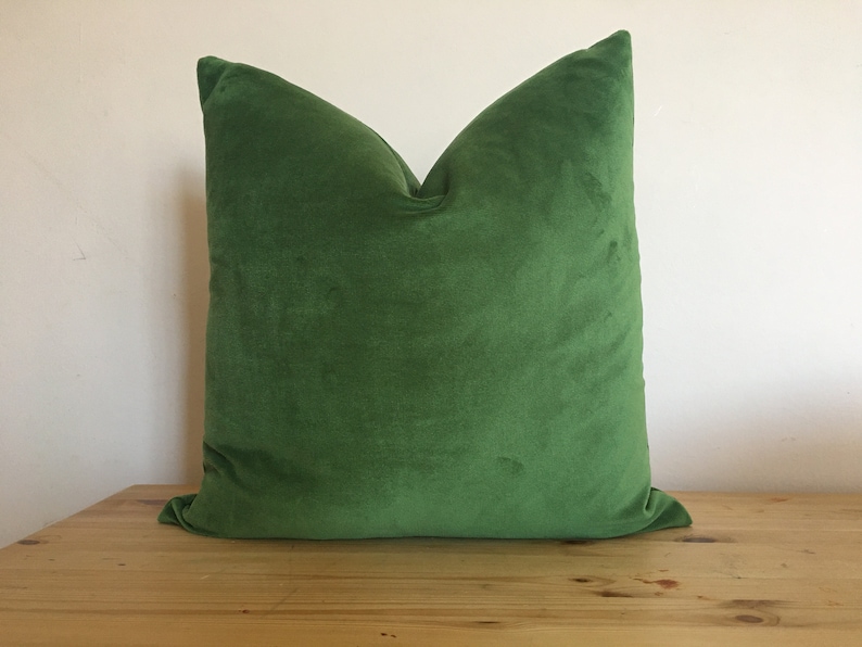 May include: A square, emerald green velvet pillow. The pillow is plush and has a soft, textured appearance. The pillow is sitting on a light brown wooden surface. Home decor item.