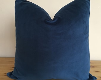 May include: A square, navy blue velvet pillow. The pillow is a solid color and has a soft, plush appearance. The pillow is resting on a wooden surface.