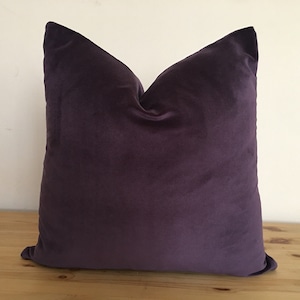 Puede incluir: Un coj&iacute;n cuadrado de color morado oscuro. El coj&iacute;n tiene una textura suave y aterciopelada y est&aacute; colocado sobre una superficie de madera. El color es uniforme, sin patrones ni dise&ntilde;os visibles. Mide aproximadamente 45 cm x 45 cm.