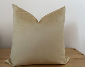 May include: A square, light beige pillow with a soft, velvety texture. The pillow is placed on a wooden surface and leans against a white wall. The pillow's color is a warm, neutral tone, suitable for home decor.