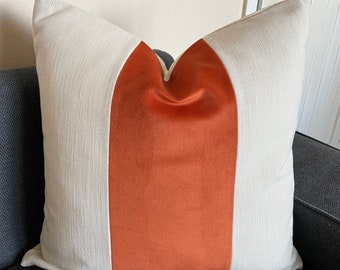 May include: Square decorative pillow with a textured off-white fabric on the sides and a central vertical panel of burnt orange velvet. The pillow is placed on a dark gray sofa, showcasing its design and color contrast.