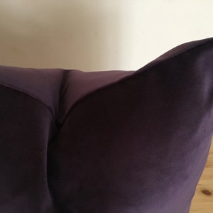 Puede incluir: Primer plano de un coj&iacute;n morado oscuro. El coj&iacute;n tiene una textura suave y aterciopelada y un brillo sutil. El coj&iacute;n es de un tono morado intenso, con una ligera variaci&oacute;n de tono en su superficie. El coj&iacute;n est&aacute; sobre una superficie de madera.