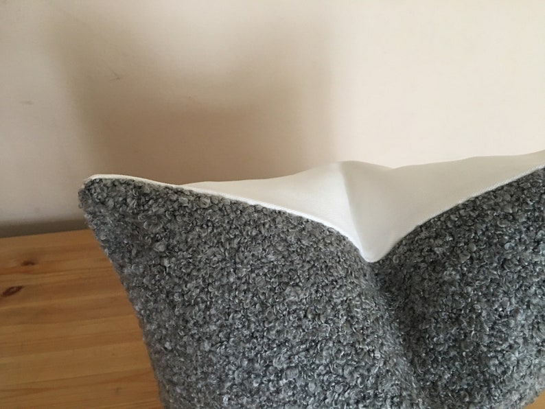 May include: A decorative pillow with a textured gray fabric on the bottom half and a smooth white fabric on the top half. The pillow is resting on a wooden surface.