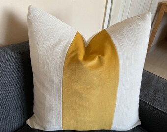 May include: Square decorative pillow with a white and mustard yellow color block design. The pillow has a textured white fabric on the sides and a smooth mustard yellow fabric in the center. The pillow is placed on a dark gray sofa.