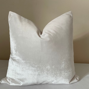 May include: A square, off-white pillow with a soft, velvety texture. The pillow has a subtle sheen and a classic, inviting shape. The neutral color makes it suitable for various decor styles. The pillow is likely filled with a soft material for comfort.