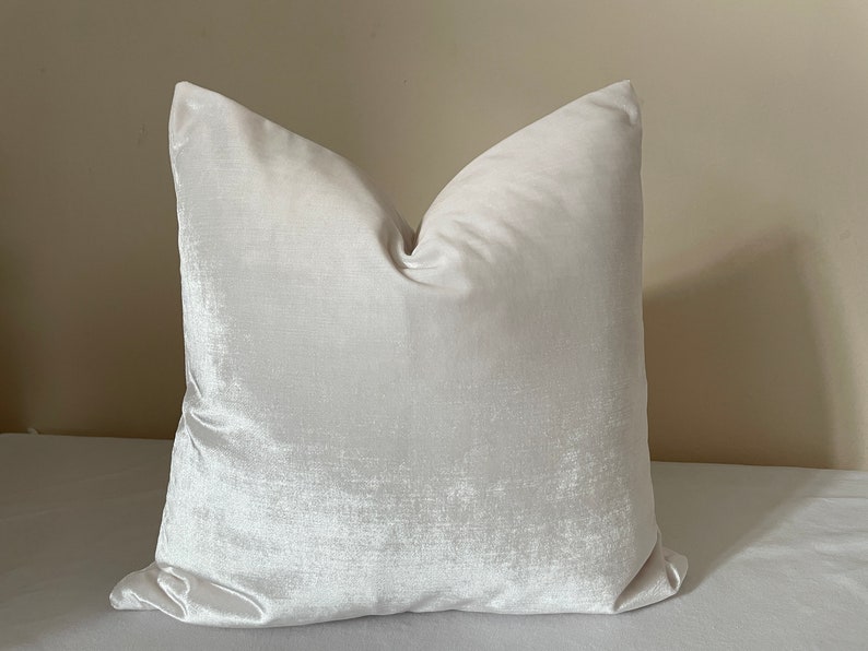 May include: A square, off-white pillow with a soft, velvety texture. The pillow has a subtle sheen and a classic, inviting shape. The neutral color makes it suitable for various decor styles. The pillow is likely filled with a soft material for comfort.