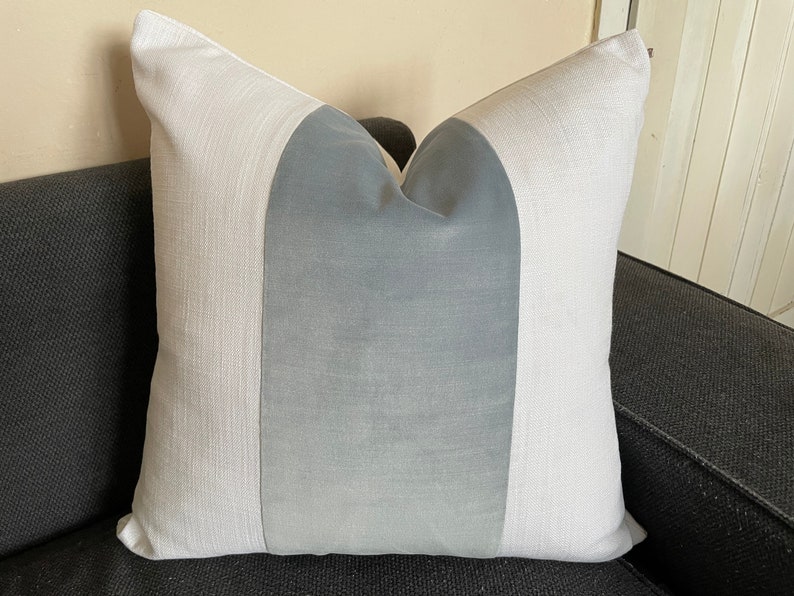 May include: A decorative square pillow with a color block design. The pillow features a central panel of light blue fabric, flanked by vertical panels of white fabric. The pillow is placed on a dark gray sofa.