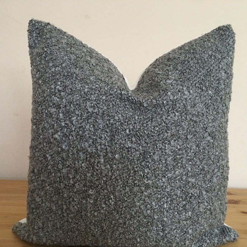 May include: A square decorative pillow with a textured, gray fabric cover. The pillow has a soft, plush appearance and is resting on a wooden surface. The pillow's neutral color and texture suggest a cozy, modern aesthetic.