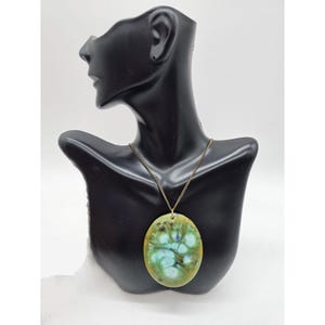 Puede incluir: Un collar con un colgante ovalado de cerámica verde y azul con una cadena dorada. El colgante tiene un diseño marmoleado con remolinos de verde, azul y blanco.