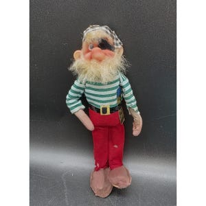 May include: A pirate doll with a tan face, a long blonde beard, and a black eye patch. The doll wears a green and white striped long-sleeve shirt, a black belt with a yellow buckle, and red trousers. It has a black and white checkered head covering.