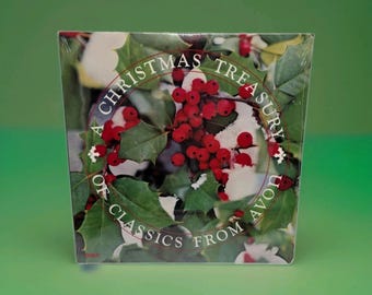 Op de afbeelding: Een vierkante albumhoes met de titel A Christmas Treasury of Classics from Avon, voorzien van een foto van felrode hulstbessen en groene bladeren bestrooid met witte sneeuw, omringd door een cirkelvormige witte rand.