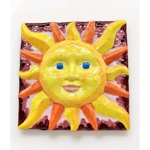 May include: A square ceramic sun with a smiling face and radiating rays. The sun is yellow with orange and yellow rays, blue eyes, and a pink mouth. The background is a dark maroon colour.