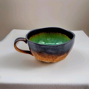 May include: A ceramic mug with a wide, shallow bowl shape and a single handle. The exterior features a textured, mottled finish transitioning from dark charcoal at the rim to a warm, sandy brown at the base. The interior is a vibrant, glossy green.