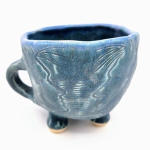 Pode incluir: Uma chávena de chá de cerâmica azul com um design texturizado e três pés. A chávena tem uma pega e é feita em estilo rústico.