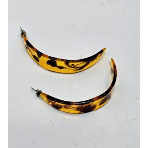 Pode incluir: Um par de brincos de argola com formato curvo, semelhante a uma banana. Os brincos apresentam um padrão de tartaruga com tons de amarelo e castanho. Os pinos são prateados.
