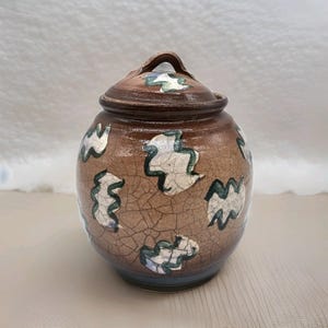 May include: A brown ceramic jar with a lid, featuring a crackle glaze and abstract white and green leaf-like designs. The jar has a rounded shape with a handle on the lid. The base has a blue band.