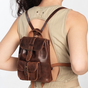 May include: A brown leather backpack with two front pockets and a buckle closure. The backpack is being worn by a person wearing a light brown sleeveless dress.