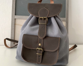 Puede incluir: Una mochila de lona gris con detalles de cuero marrón y cierre de hebilla. La mochila tiene un pequeño bolsillo frontal con cierre de hebilla.