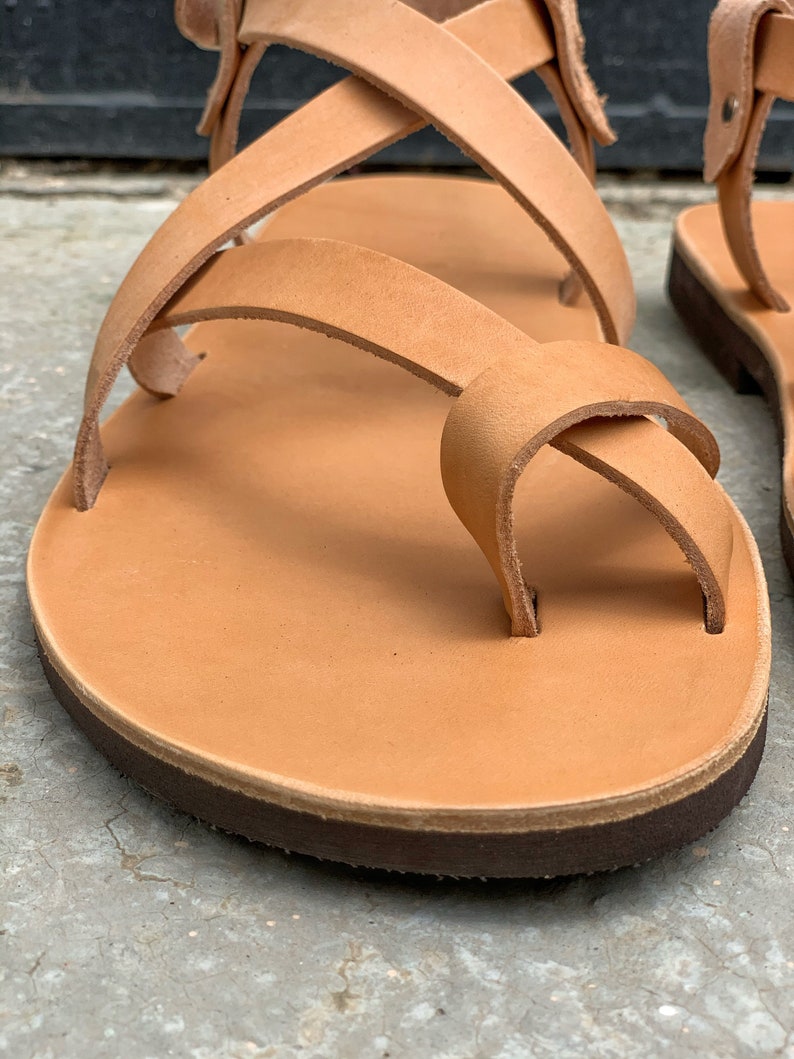 Pu&ograve; includere: Sandali in pelle color cuoio con pi&ugrave; cinturini che si incrociano sul piede. I sandali hanno una suola in pelle marrone.