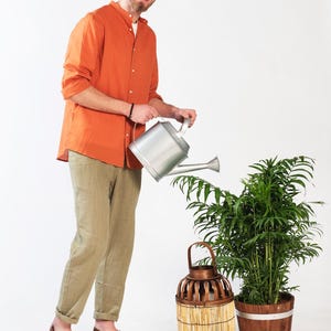 Puede incluir: Un hombre con una camisa naranja y pantalones caqui est&aacute; regando una planta en maceta con una regadera plateada. Una linterna de mimbre marr&oacute;n est&aacute; al lado de la planta.