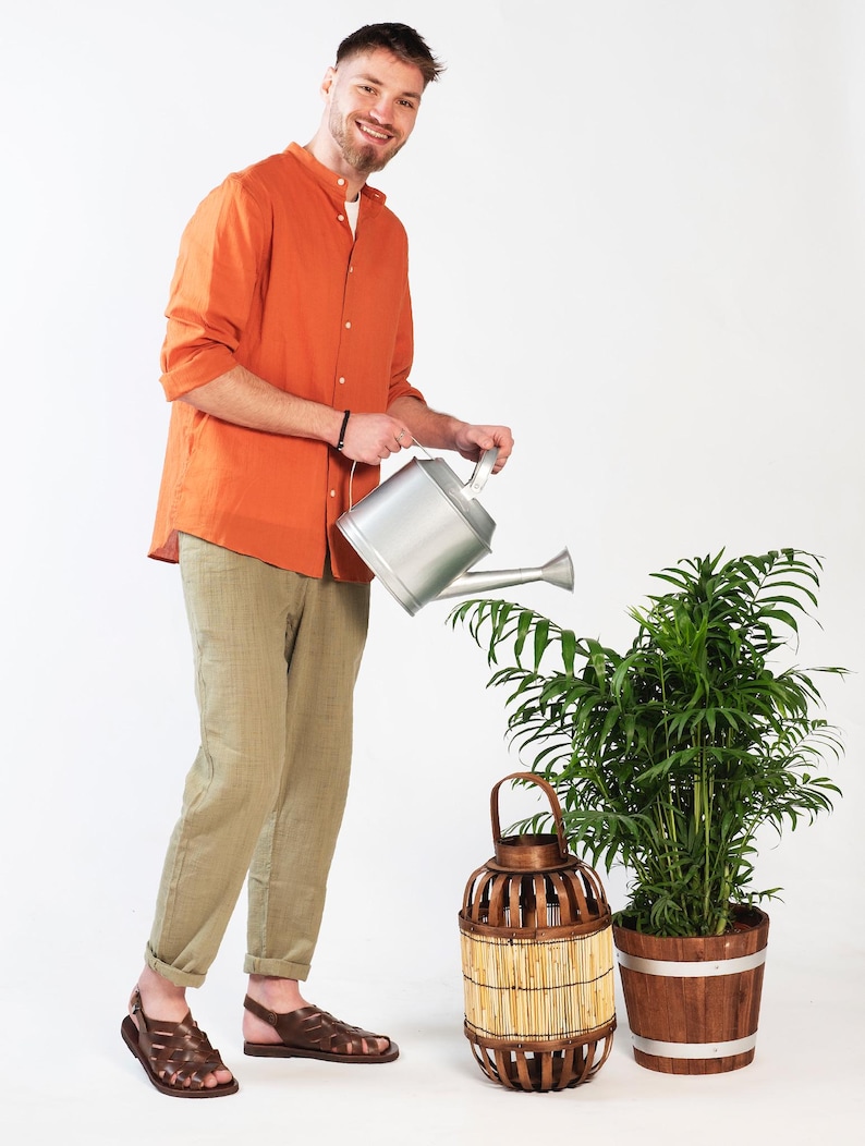 Puede incluir: Un hombre con una camisa naranja y pantalones caqui est&aacute; regando una planta en maceta con una regadera plateada. Una linterna de mimbre marr&oacute;n est&aacute; al lado de la planta.