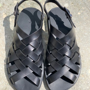 Puede incluir: Un par de sandalias de cuero negro con un dise&ntilde;o tejido. Las sandalias tienen un cierre de hebilla y una suela plana.