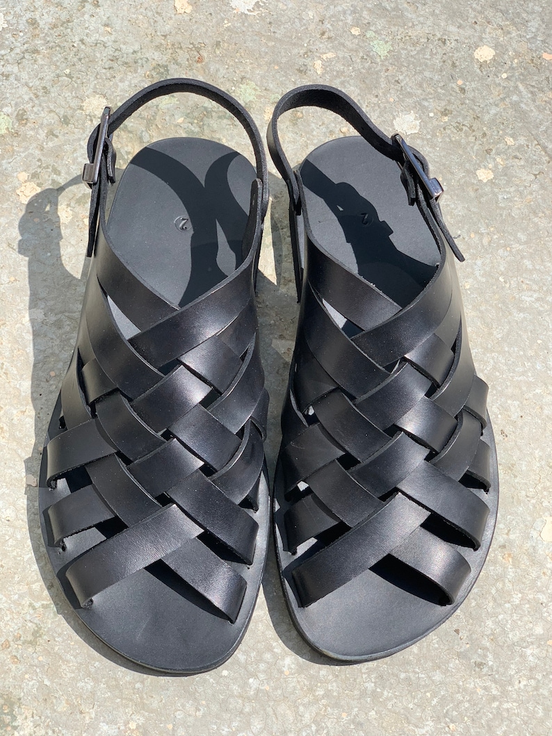 Puede incluir: Un par de sandalias de cuero negro con un dise&ntilde;o tejido. Las sandalias tienen un cierre de hebilla y una suela plana.