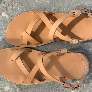 May include: A pair of tan leather sandals with criss-cross straps and a buckle closure. The sandals are size 5.