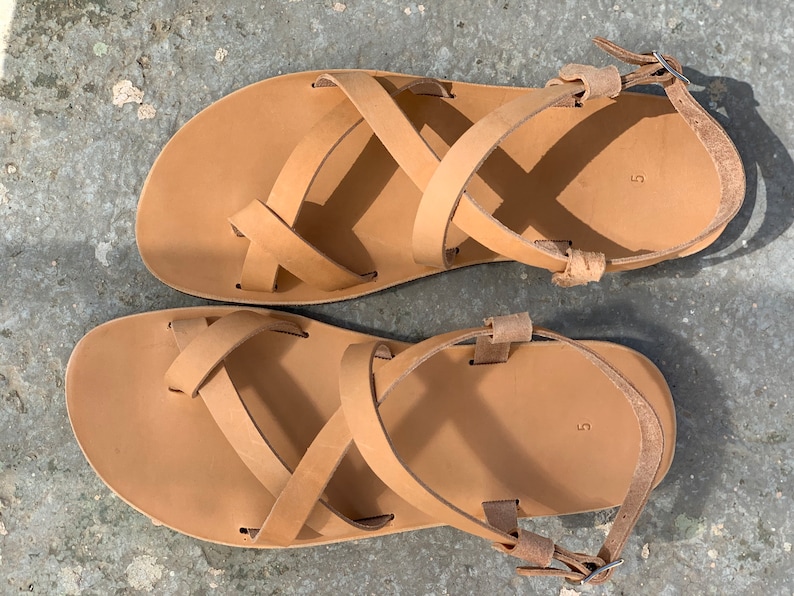 May include: A pair of tan leather sandals with criss-cross straps and a buckle closure. The sandals are size 5.