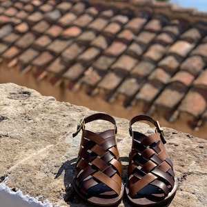 Puede incluir: Un par de sandalias de cuero marr&oacute;n con un dise&ntilde;o tejido. Las sandalias tienen un cierre de hebilla y una suela plana.