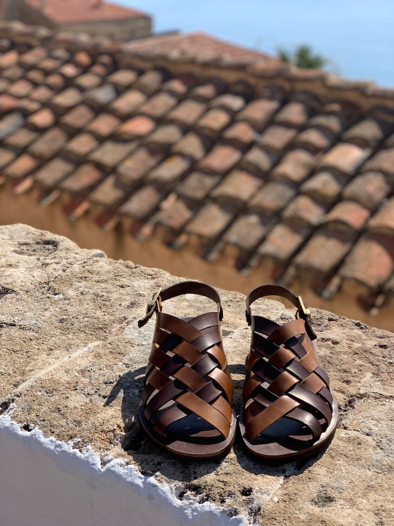 Puede incluir: Un par de sandalias de cuero marr&oacute;n con un dise&ntilde;o tejido. Las sandalias tienen un cierre de hebilla y una suela plana.