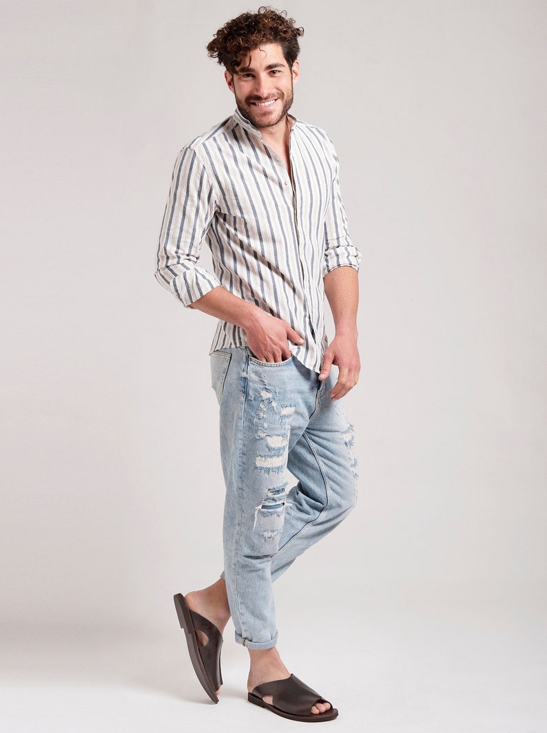 May include: A man wearing a striped button-down shirt, light blue ripped jeans, and brown leather sandals. The shirt has vertical stripes in white and blue. The jeans are distressed with multiple rips. The sandals are open-toed.