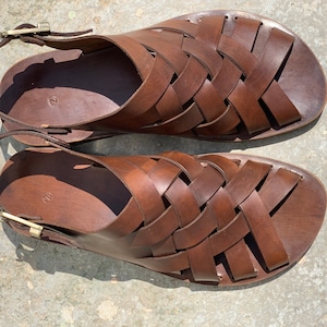 Puede incluir: Un par de sandalias de cuero marr&oacute;n con un dise&ntilde;o tejido. Las sandalias tienen un cierre de hebilla en la correa del tobillo.