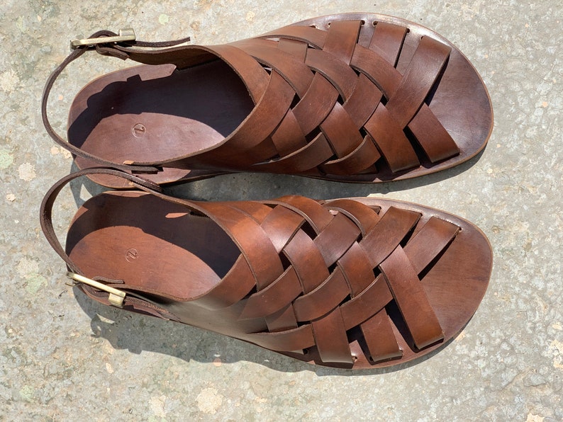 Puede incluir: Un par de sandalias de cuero marr&oacute;n con un dise&ntilde;o tejido. Las sandalias tienen un cierre de hebilla en la correa del tobillo.